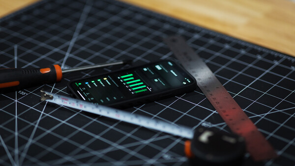 Electronic device with illuminated green LED display sits on a black cutting mat next to precision tools.
