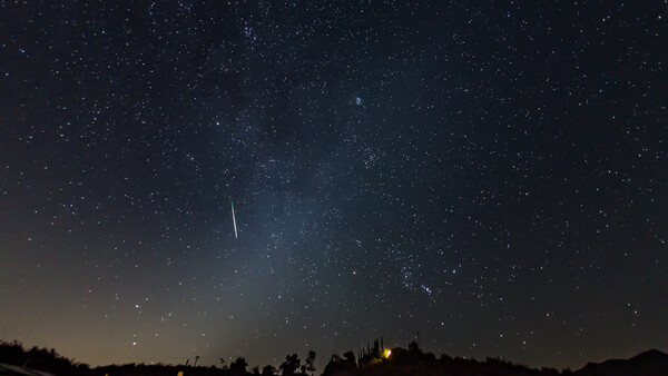 Meteor streaking across starry night sky above silhouetted landscape with warm lights below.