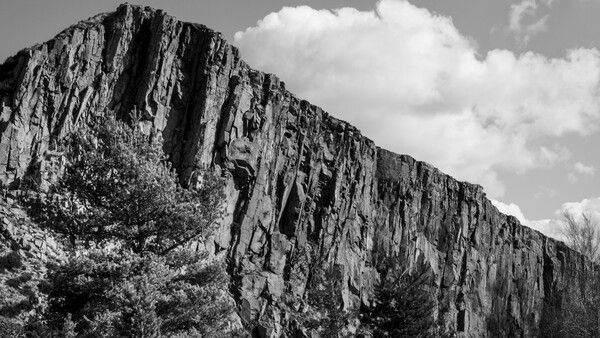 Dramatic columnar basalt cliff face rises against a cloudy sky, with vegetation at its base.