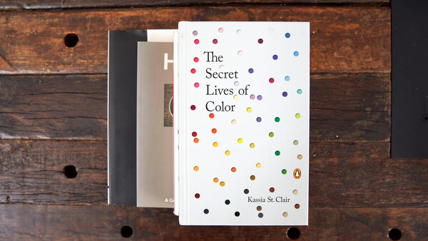 Stack of three books on wooden surface, with 'The Secret Lives of Color' by Kassia St. Clair prominently displayed on top featuring colorful dots.