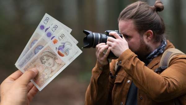 Photographer holding camera while another person displays British banknotes in front of the lens.