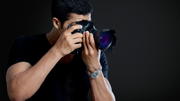 Male photographer holding DSLR camera up to his face, shooting against dark background.
