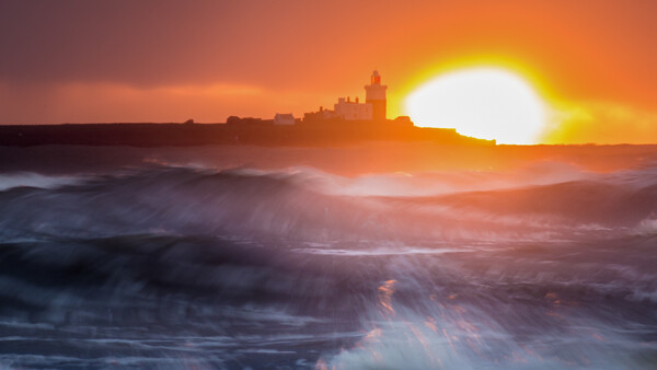 Coastal city skyline silhouetted against a bright sun setting over turbulent ocean waves.