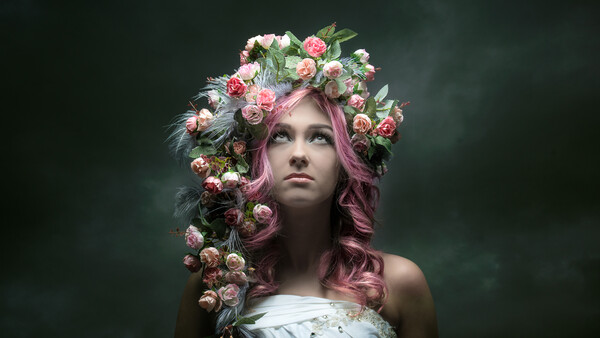 Woman with long pink hair wearing an elaborate floral crown of pink and white roses against a dark green background.