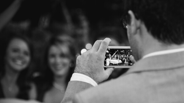 Man holding a smartphone capturing a photo of two smiling women in the background.