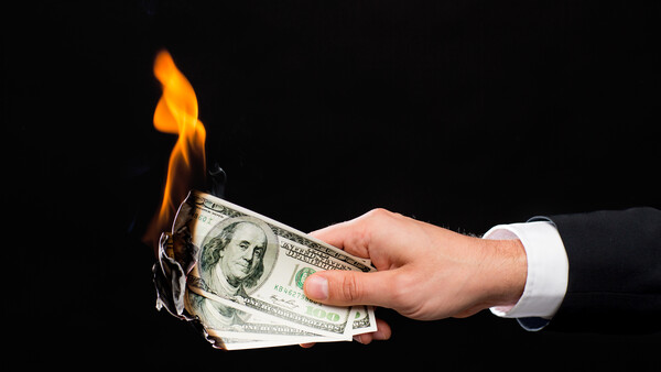 Hand in white shirt cuff holding burning U.S. dollar bills against black background.