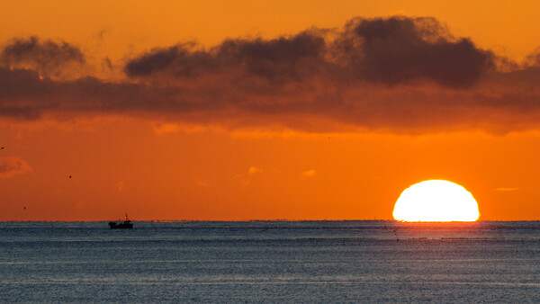 Large sun setting over calm ocean waters with a small fishing vessel silhouetted on the horizon beneath dramatic orange and brown clouds.