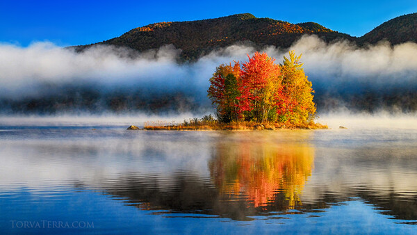 Small forested island with vibrant autumn foliage reflected in still water, surrounded by mist-covered mountains.