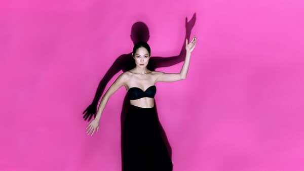 Woman in black crop top and skirt posed against bright magenta background with flowing hair and hand gestures.