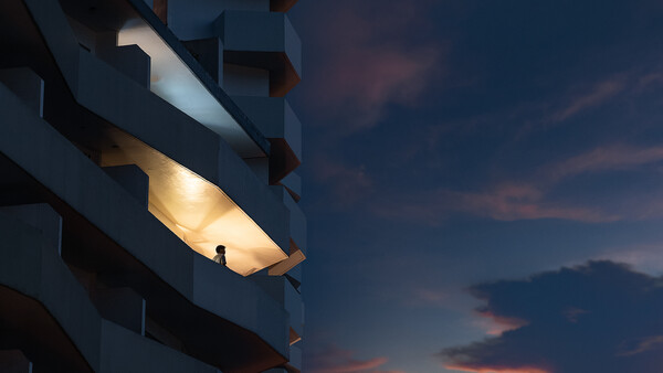 Silhouetted figure standing in an illuminated balcony alcove of a modern building at dusk.