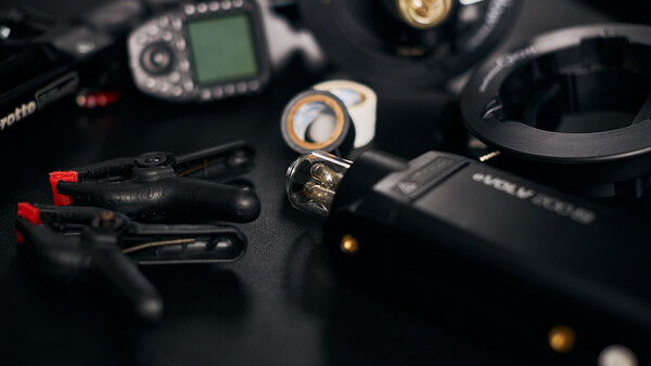 Photography and audio gear arranged on black surface including camera flash, wireless microphone, headphones, and camera body with LCD screen.