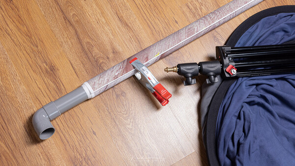 Tripod arm with red clamp and mounting hardware laid out on wooden floor next to camera bag.