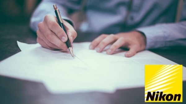 Person in light blue shirt writing on white paper with a pen at a desk.