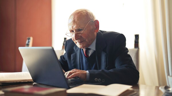 Older man wearing glasses and dark blazer working on laptop at desk in warm-lit interior space.