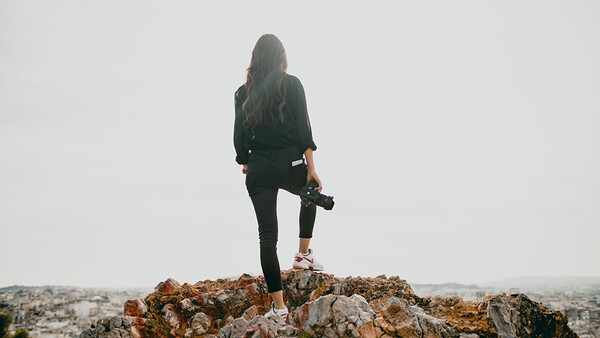 Woman standing on rocky outcrop holding camera, wearing dark clothing against overcast sky.