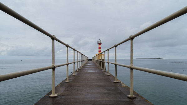 Long wooden pier extending into calm water toward a red and white striped lighthouse in the distance.