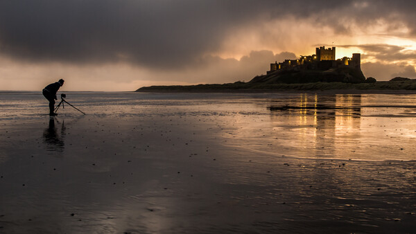 Photographer with tripod on wet beach at sunset, medieval castle ruins visible across the water.