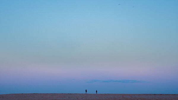 Two silhouetted figures standing on a vast, flat beach during twilight with a purple and blue sky.
