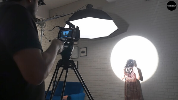 Filmmaker operating camera on tripod while subject stands before large circular backlit panel in studio.