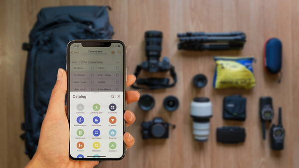 Photographer's hand holding smartphone displaying a catalog app interface with various photography equipment laid out on wooden surface.