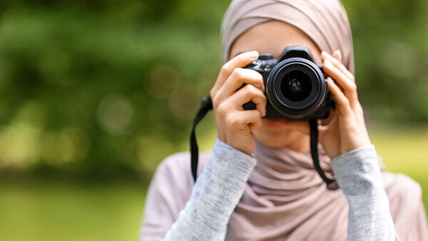 Young photographer wearing hijab holding DSLR camera with telephoto lens to face outdoors.