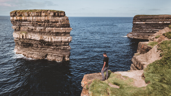 Person standing on grassy cliff edge between two large rock formations jutting from the sea.