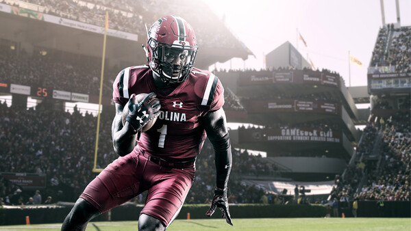Football player in maroon uniform running with ball in stadium during game.