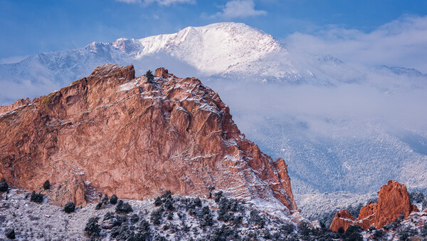 Snow-dusted red rock formation with snow-capped mountain peak in distant background under blue sky.