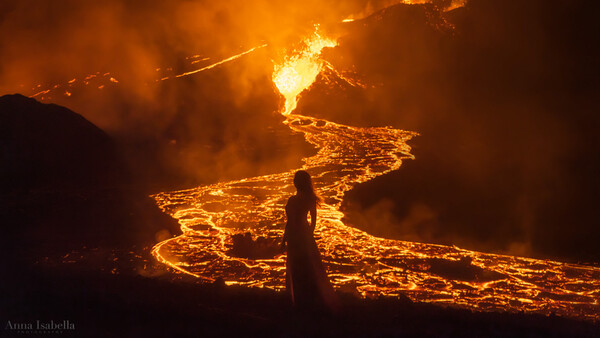 Silhouetted figure standing before flowing lava from an active volcanic eruption at night.