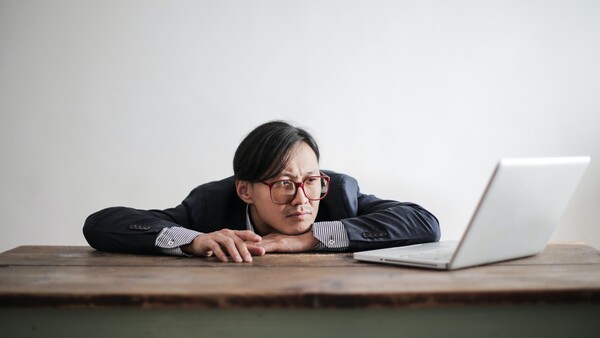 Man in business casual attire leaning on desk toward laptop with relaxed posture.
