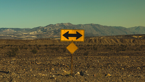 Yellow road sign with opposing arrows centered in an arid desert landscape with mountains in the distance.