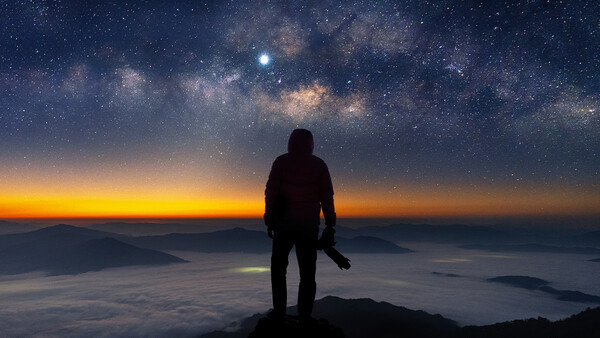 Silhouetted figure standing on a mountain peak overlooking a sea of clouds at sunset with the Milky Way visible above.
