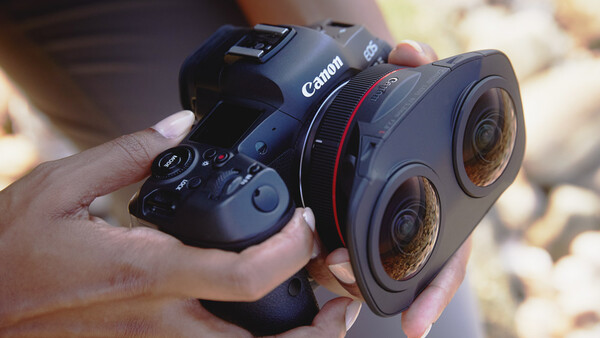 Canon camera with dual-lens attachment held in someone's hands alongside a wireless controller.