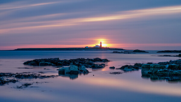 Lighthouse on distant island at sunset with rocky foreground and calm water.