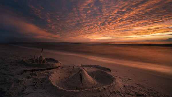 Sandcastle on a beach at sunset with dramatic orange and amber clouds overhead.