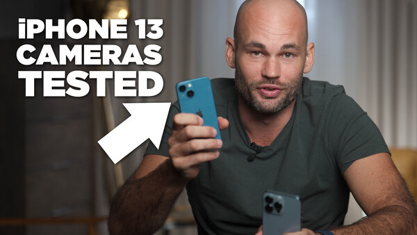 Man in dark t-shirt holding blue iPhone 13 toward camera with second device visible on table.