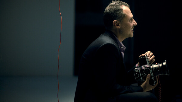 Man in dark clothing holding a film camera, photographed in profile against a minimalist dark background.