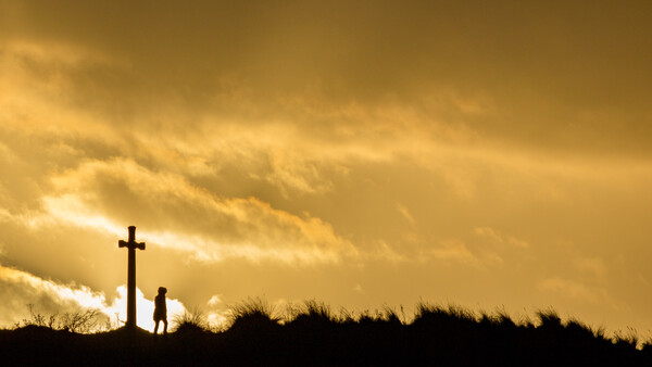 Silhouetted figure standing beside a cross on a hilltop against a golden sunset sky.