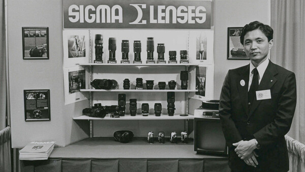Man in business attire standing beside a display shelf of Sigma lenses and camera equipment.