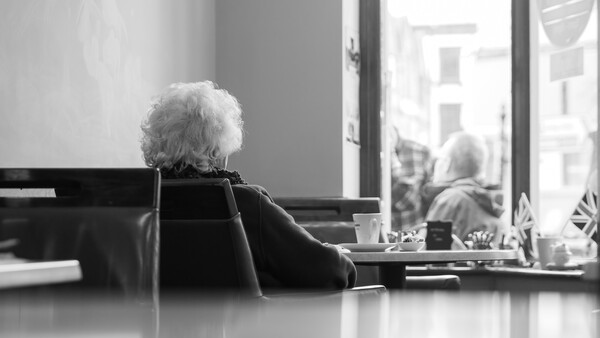 Elderly person with white hair seated at a desk, gazing out a window onto a city street.