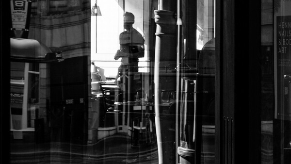Black and white street photograph showing a figure visible through glass storefront reflections and layers of windows.