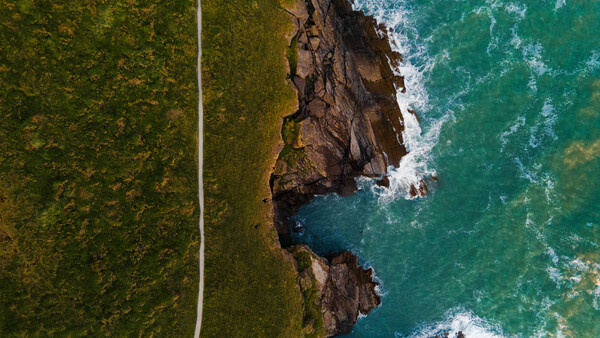 Aerial view of rocky coastline with turquoise ocean waves, dark cliffs, and dense green forest with a white road cutting through.