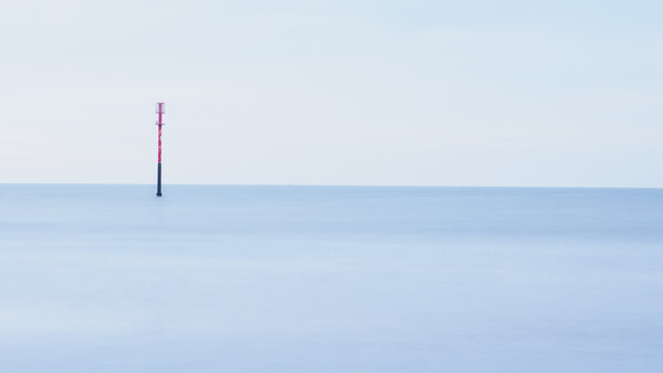 A single red and black pole stands isolated in calm, shallow blue water against a clear horizon.