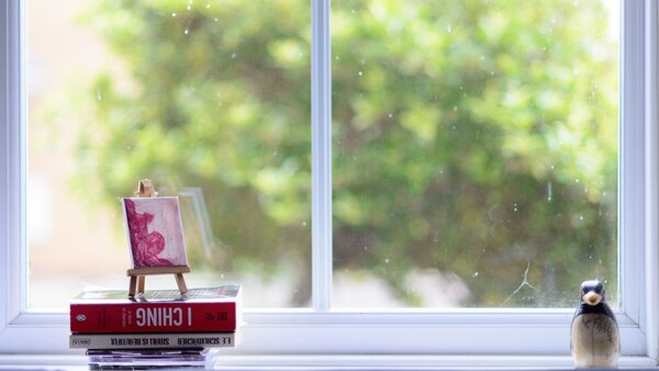 Windowsill still life with stacked books, miniature easel painting, and decorative bird figurine.