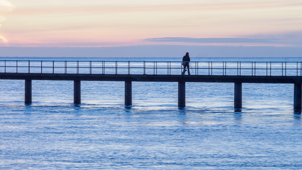 Solitary figure standing on a long pier extending into calm blue water at twilight.