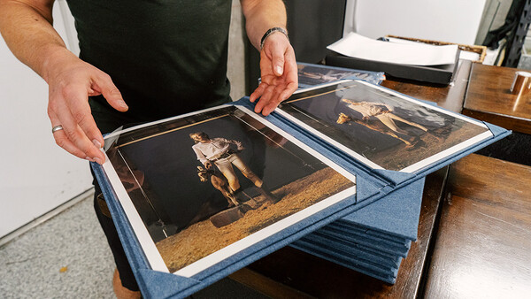 Hands holding open a blue cloth portfolio displaying two vintage black and gold photographs.