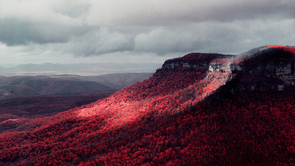 Mountainous landscape with dense red autumn foliage beneath dramatic cloudy sky.