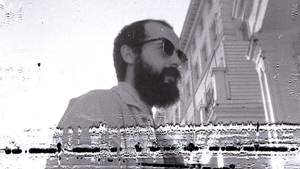 Bearded man wearing sunglasses and light-colored shirt, photographed in profile against urban staircase backdrop.