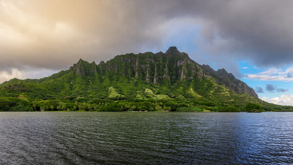 Lush green mountain ridge rising from calm blue water under partly cloudy sky.