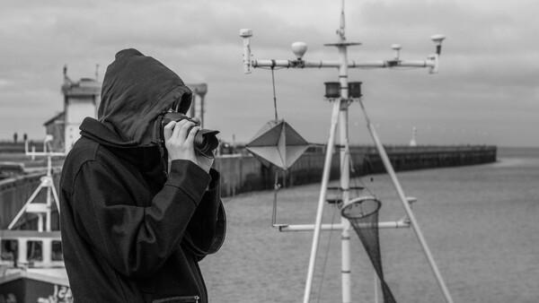 Photographer in black hoodie holding camera, shooting from a research vessel with scientific equipment visible in background.